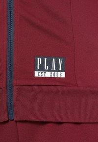Bordeauxfarbene Zip-Jacke mit marineblauen Akzenten, die auf der Vorderseite ein schwarz-weißes Label mit der Aufschrift "PLAY EST. 2006" deutlich präsentiert. Glatte Stoffoberfläche.