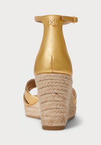 Zlatne kožne sandale na wedge potpeticu s tkanim jutenim detaljima, remenom oko gležnja i RL logotipom na stražnjem dijelu. Teksturirana potpetica dodaje detalje dizajnu.