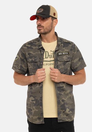 Homme portant un t-shirt beige Von Dutch sous une chemise camouflage déboutonnée et une casquette de baseball bicolore Von Dutch, regardant vers la gauche.