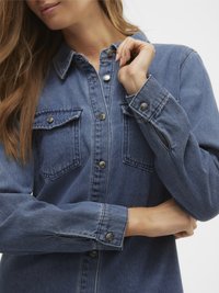 Džinsinis marškinėlis vidutiniškai mėlynos spalvos su spaustukais, dviem krūtinės kišenėmis ir apykakle; ilgomis rankovėmis su sagų apvadais ir minkštu audiniu.