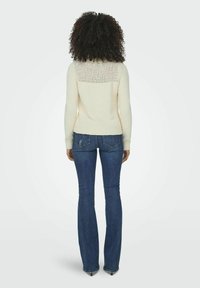 Pull en mailles crème avec un dos texturé, présentant des manches longues et un ourlet côtelé. Associé à un jean bleu foncé ajusté et des talons noirs pointus.