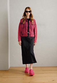Rosa denimjakke over en svart topp, kombinert med et langt svart skjørt og rosa clogs. Glatte solbriller fullfører antrekket.