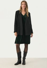 Schwarzer doppelt geknöpfter Blazer über einem knielangen, dunkelgrünen Samtkleid, kombiniert mit schwarzen Leder-Cowboy-Stiefeln, glatte Textur.