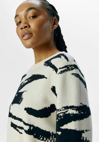 Strikket sweater i creme med iøjnefaldende sorte abstrakte mønstre. Blød tekstur, runde halsudskæring og let sænkede skuldersømme for komfort.