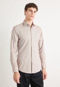 Lys beige langermet skjorte med knappelukking foran og spiss krage, laget av glatt stoff. Har figursydd design og buet nederkant.
