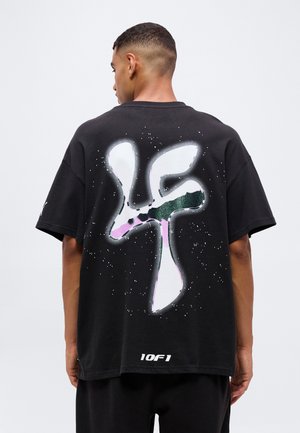 Mężczyzna widziany od tyłu, noszący czarny oversize T-shirt z dużą białą i zieloną abstrakcyjną grafiką oraz tekstem "10F1" na dole pleców.