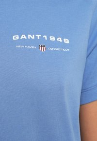 Kék pamut póló fehér "GANT1949" felirattal és pajzs emblémával. Kerek nyakú, rövid ujjú, sima textúrájú.