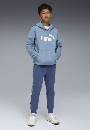 Sudadera azul claro con el logotipo blanco de "PUMA", bolsillo frontal, combinada con pantalones de chándal azul marino que tienen un gráfico lateral. Los zapatos son blancos con suelas marrones.