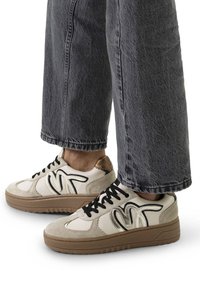 Beige Sneakers mit schwarzen Akzenten und goldenen Details, ausgestattet mit einem strukturierten Obermaterial aus Wildleder und Leder, dicker brauner Gummisohle und traditionellen Schnürsenkeln.