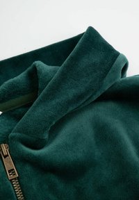 Ciemnozielony materiał polarowy o miękkiej fakturze, złożony kołnierz i widoczny mosiężny zamek po jednej stronie.