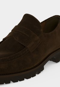 Mocassin en daim marron foncé avec semelle en caoutchouc noire épaisse et détail de bride sur le cou-de-pied sur fond clair uni.