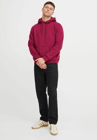 Sudadera burdeos con cordones, bolsillo tipo canguro y puños acanalados, combinada con pantalones negros y zapatillas blancas con detalles en marrón.