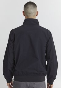 Giacca a vento blu navy con colletto alto, polsini e vita elasticizzati. Tessuto testurizzato con finitura liscia e design minimalista.