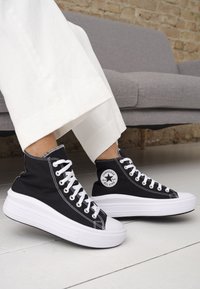 Sneakers alti in canvas nero con lacci bianchi, punta in gomma e una spessa suola piatta bianca. Presentano una toppa rotonda con logo sul lato.