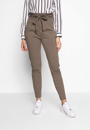 Kobieta ubrana w spodnie typu paperbag w kolorze taupe z paskiem do wiązania, bluzkę w paski z długim rękawem oraz białe trampki, stojąca na białym tle.