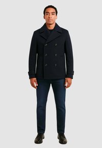 Cappotto doppiopetto navy con grandi bottoni neri, tasche laterali e un ampio colletto, abbinato a jeans blu scuro e scarpe nere.