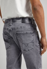 Jeans de mezclilla gris con un acabado texturizado, que cuentan con dos bolsillos traseros con solapas angulares y un parche de cuero con marca en la cintura.