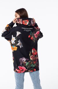 Sudadera negra con un patrón floral en colores vibrantes, que cuenta con una capucha amplia y texto en la parte posterior. El material parece suave y texturizado.