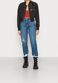 Blouson aviateur noir avec poche zippée, haut côtelé rouille, jean bleu clair déchiré et bottes noires épaisses à boucle.