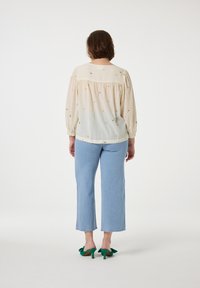 Bestickte cremefarbene Bluse mit gerafften Ärmeln, kombiniert mit hellblauen, knöchelfreien Jeans und Schuhen mit grünen Schleifenakzenten. RÜCKANSICHT.