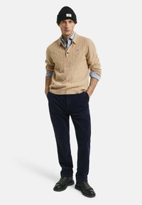 Man in een zwarte beanie, beige kabelgebreide trui over een blauwe shirt, marineblauwe corduroy broek en zwarte veterschoenen, staand met zijn handen in zijn zakken.