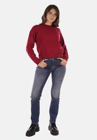 Maglione rosso a maglia con orlo a coste, abbinato a jeans blu e stivaletti neri con lacci. Il modello si trova davanti a uno sfondo bianco.
