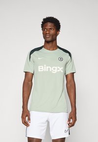 Homme portant un maillot d'entraînement léger vert du Chelsea Football Club avec le logo du sponsor Bingx et un short Nike blanc, se tenant devant un fond uni.