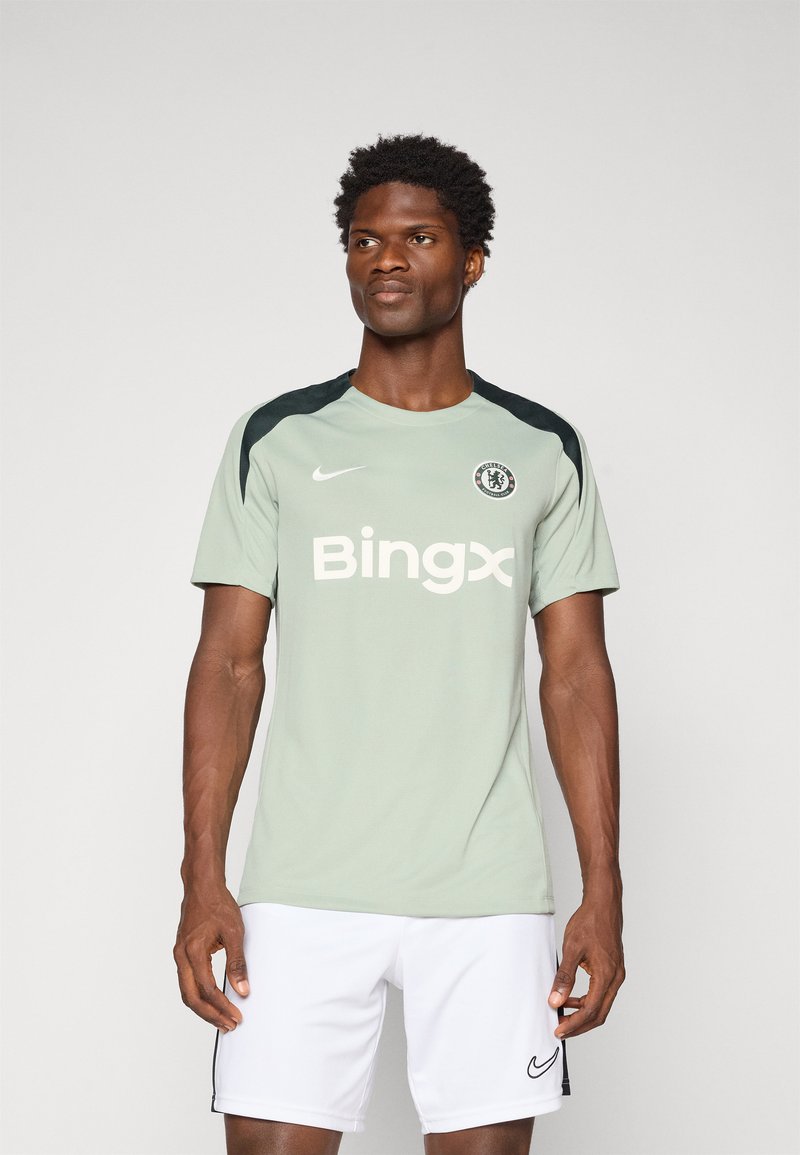 Homme portant un maillot d'entraînement léger vert du Chelsea Football Club avec le logo du sponsor Bingx et un short Nike blanc, se tenant devant un fond uni.