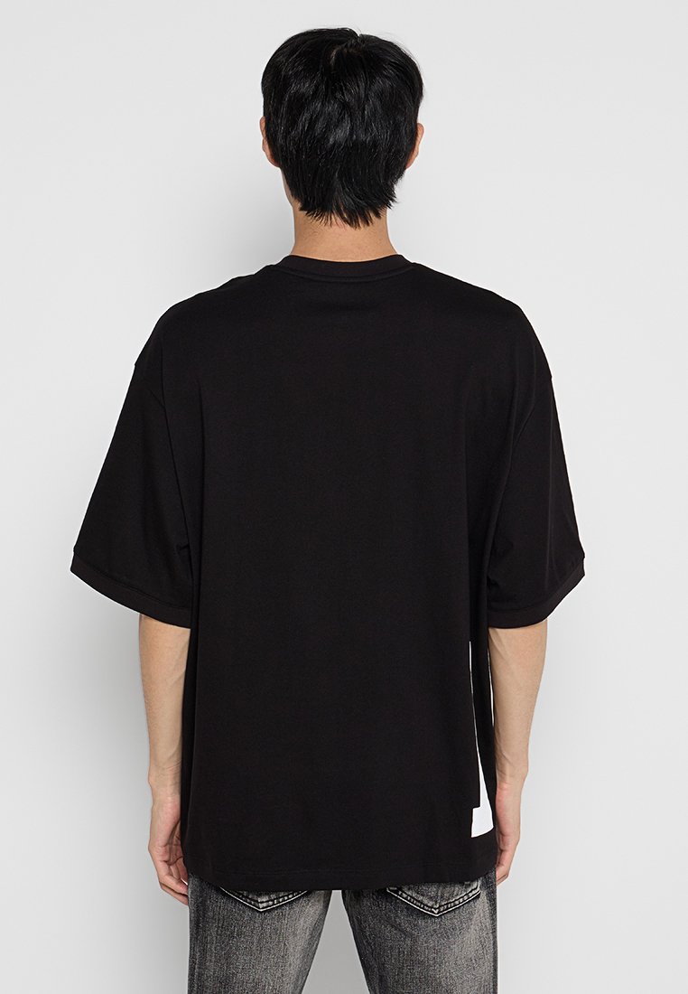 Black oversized T-shirt with short sleeves, soft fabric, and a round neck. Features a white label accent at the bottom left side.