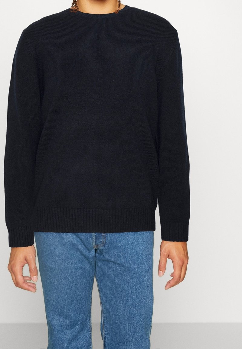 Pull en tricot bleu foncé avec un col rond côtelé et des poignets, associé à un jean bleu clair. Texture lisse, coupe décontractée, couleur unie.