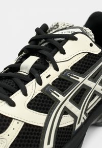 Suurendatud must-valge spordijalats, millel on võrgukangas, mustad paelad ja silmatorkav Asicsi logo küljel.