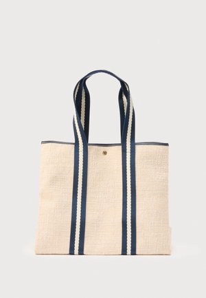 Sac cabas beige tissé, avec de longues anses bleu marine à rayures crème et un petit emblème doré, sur un fond uni.