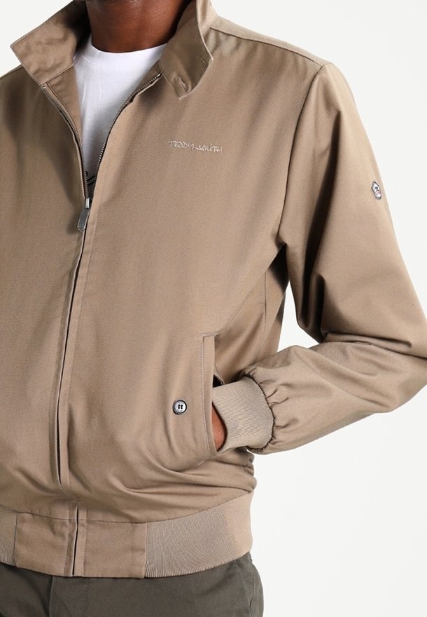 Veste beige zippée avec col montant et poignets côtelés, portée sur une chemise blanche, avec un petit logo sur la poitrine et une main dans la poche.
