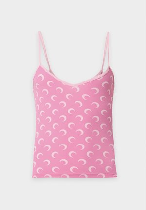 Vrouw draagt een roze spaghetti top met halve maan print en een wijd uitlopende blauwe spijkerbroek, met één hand in de zak, tegen een effen achtergrond.
