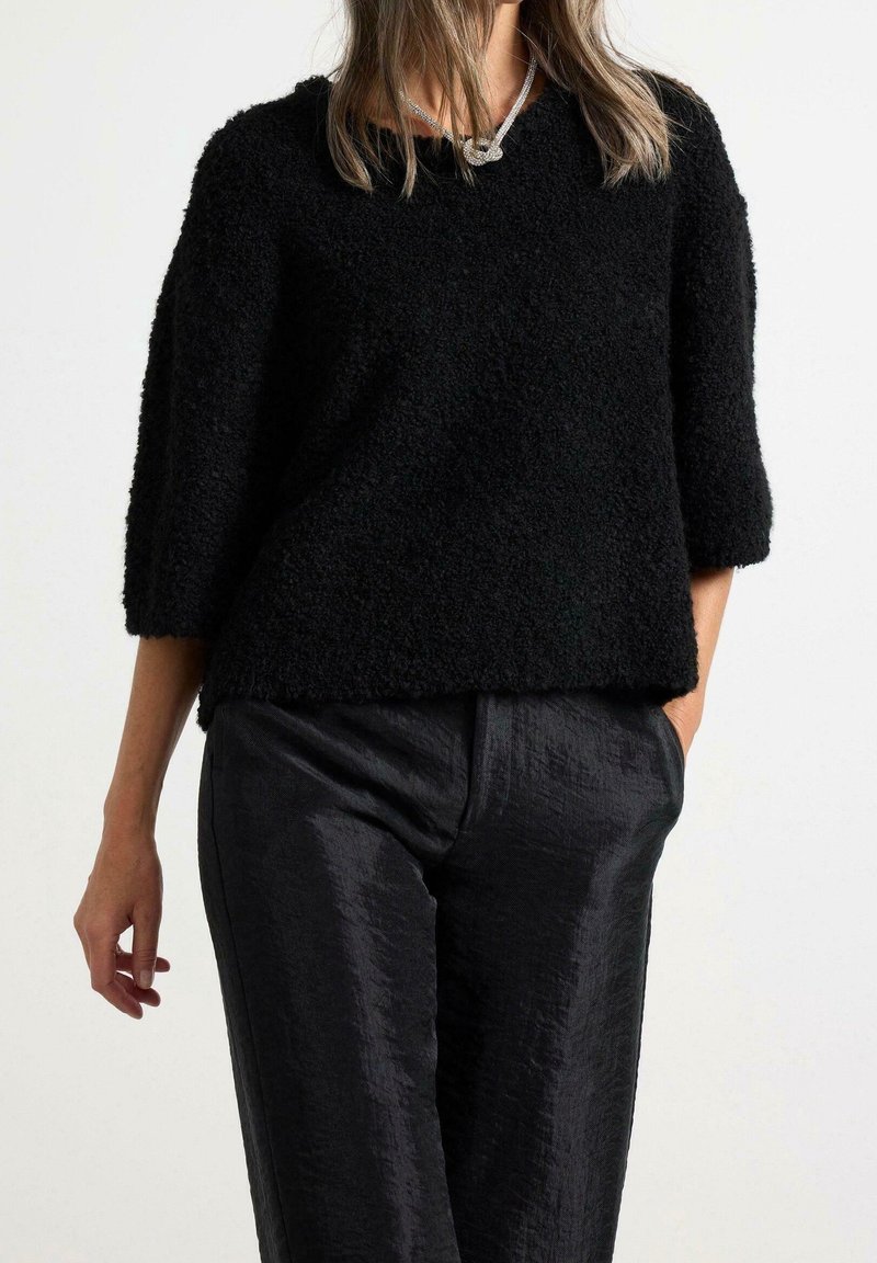 Mustasta, tekstuurillisesta neulepaidasta, jossa on rento leikkaus ja kolmanneksen mittaiset hihat, yhdistettynä kiiltäviin mustiin housuihin. Yksinkertainen muotoilu ja pehmeä materiaali.