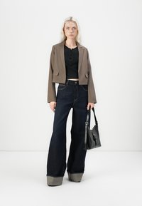 Bruine cropped blazer, zwarte button-up top, wide-leg donkerblauwe jeans met grijze manchetten en een zwarte schoudertas. Minimalistisch ontwerp en texturen.