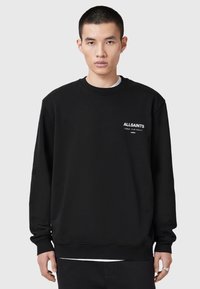 Young man with short hair wearing a black AllSaints sweatshirt with white text on left chest, standing against a plain gray background.