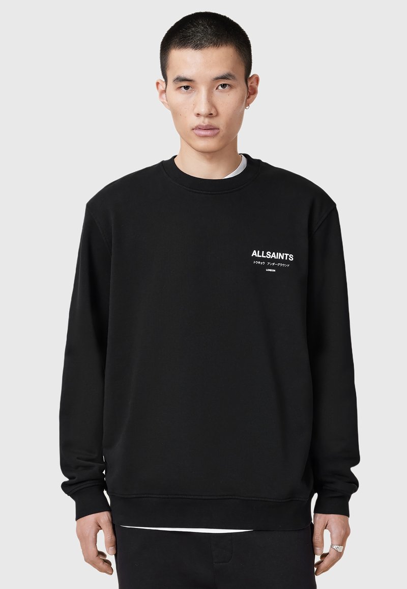 Young man with short hair wearing a black AllSaints sweatshirt with white text on left chest, standing against a plain gray background.