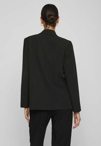 Černý blejzr s strukturovaným designem, dlouhými rukávy a rovný střihem. Vyznačuje se jemným leskem a texturovanou látkou, nošený přes kalhoty ze stejného materiálu.