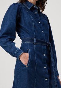 Jeansklänning i mörkblått, med krage, knapprad framtill, belagd midja och sidofickor med kontrasterande sömmar.