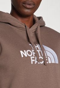 Sweatshirt castanho feito de tecido macio, apresentando um design de logotipo branco e azul claro. Inclui cordões e um clássico bolso tipo canguru.