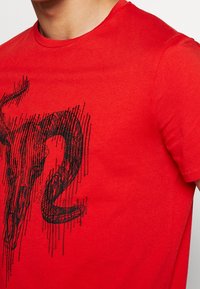 Červené bavlněné tričko s černým grafickým designem lebky a abstraktních linií na přední straně. Kulatý výstřih a krátké rukávy.