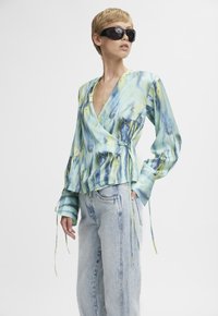 Blusa avvolgente in seta con un vivace motivo ad acquerello blu e verde, maniche lunghe e lacci in vita, abbinata a jeans in denim blu chiaro.