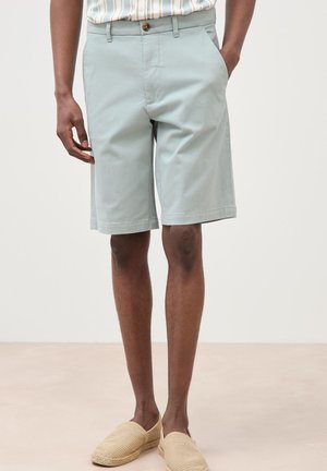 Mittellange hellblaue Shorts, getragen mit einem gestreiften Hemd, das hineingesteckt ist, kombiniert mit beigen gewebten Slip-On-Schuhen bei einer stehenden Person.