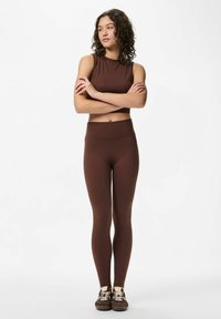 Smeđi set aktivne odjeće koji sadrži majicu bez rukava i tajice visokog struka, izrađen od rastezljivog, glatkog materijala. Ležerna obuća upotpunjava izgled.