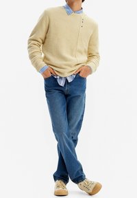 Pull en polaire couleur crème avec détails de boutons sur les côtés, superposé à une chemise rayée bleu clair. Porté avec un jean bleu et des baskets crème.