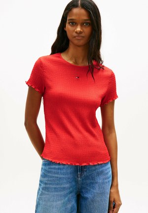 Vrouw draagt een nauwsluitend rood shirt met korte mouwen en gerimpelde randen, gecombineerd met een blauwe spijkerbroek, staand tegen een effen witte achtergrond.