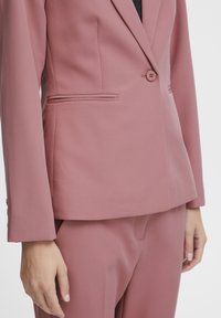 Vaaleanpunainen räätälöity bleiseri, jossa on yksirivinen nappi, lovikaulus ja sivutaskut. Kangas on sileää ja mattapintaista. Yhdistetty samansävyisiin housuihin.