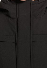 Veste à capuche noire à texture lisse, dotée de deux grandes poches avant et d'un design minimaliste. Cordon de serrage à la capuche pour ajustement.