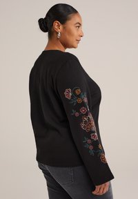 Kvinde med h�året i en knold iført en sort langærmet top med farverig blomsterbroderi på ærmet og mørkegrå jeans.